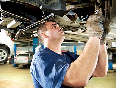 Mechanic in blue coveralls inspecting the undercarriage of a hoisted vehicle.